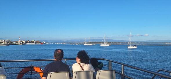 The photo shows two adults and one child on the upper deck of the electric ferry that connects São Jacinto (Aveiro) and Forte da Barra (Ílhavo). In the background: the São Jacinto  harbor and waterfront.