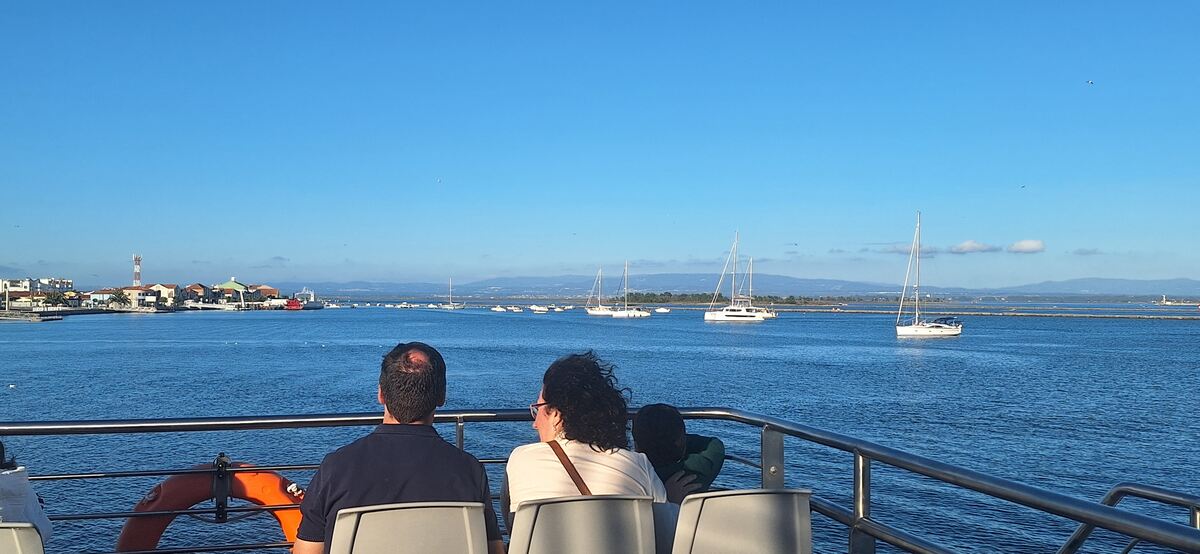 The photo shows two adults and one child on the upper deck of the electric ferry that connects São Jacinto (Aveiro) and Forte da Barra (Ílhavo). In the background: the São Jacinto  harbor and waterfront.